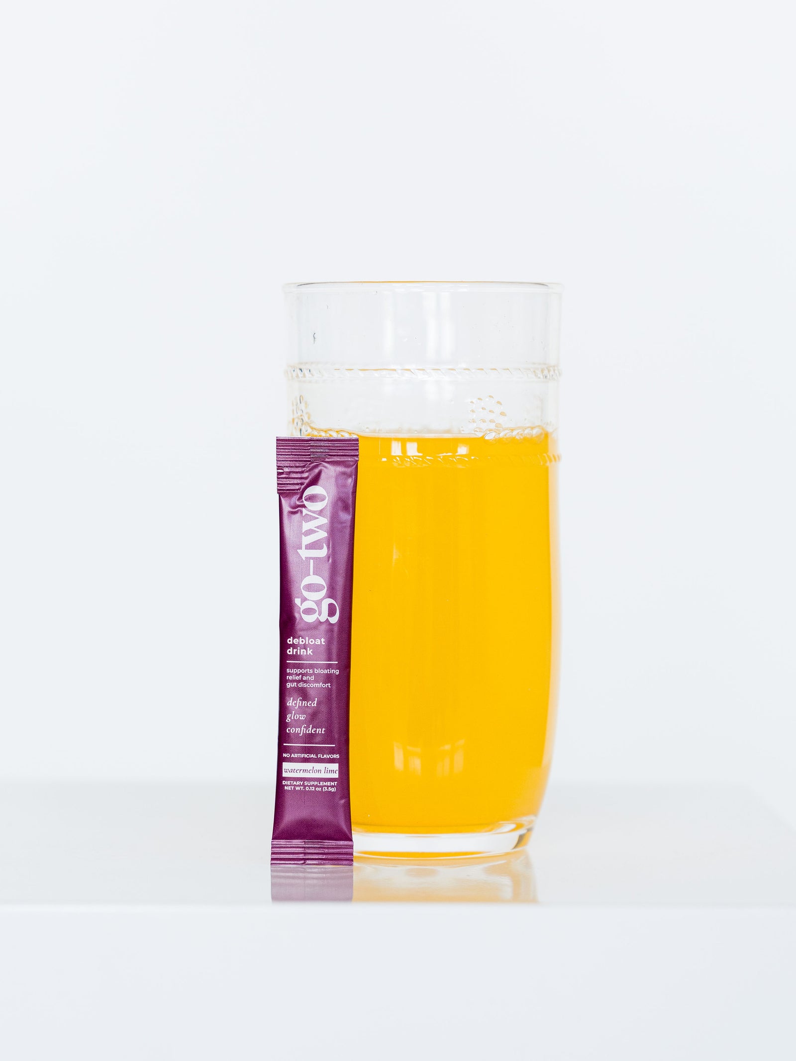 Go-Two Debloat stick pack beside a glass of watermelon lime drink on a white pedestal