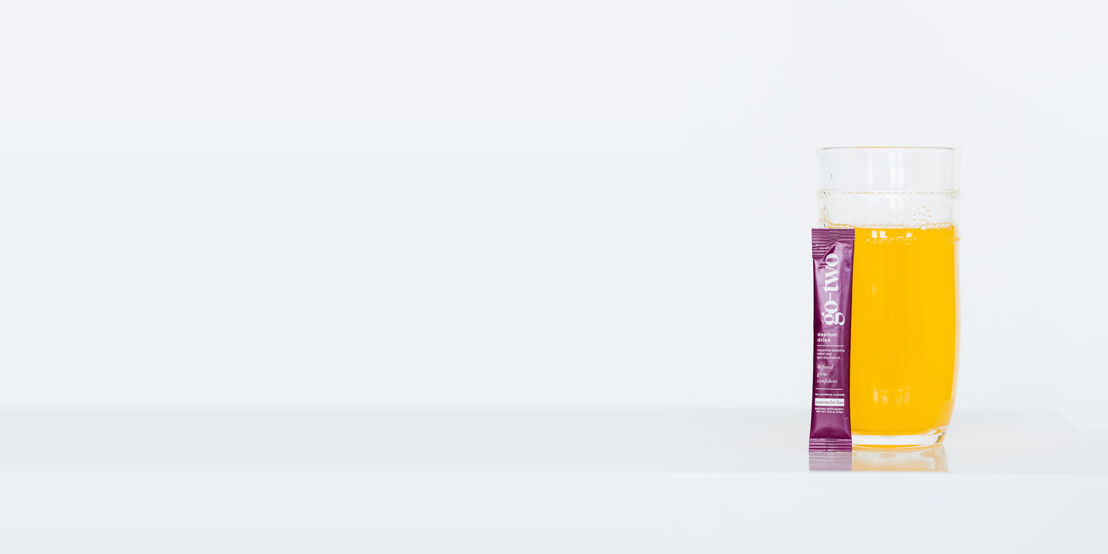 A Go-Two Debloat Watermelon Lime stick pack standing beside a tall glass of the prepared drink on a white surface